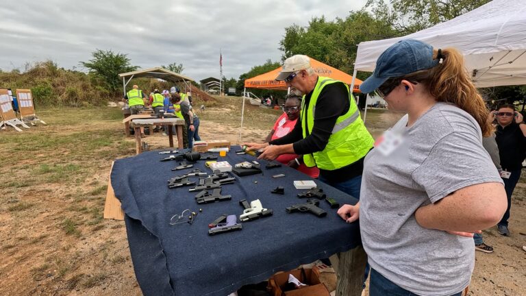 A club instructor demonstrates firearm maintenance to participants at a shooting station, with other stations and staff visible behind.