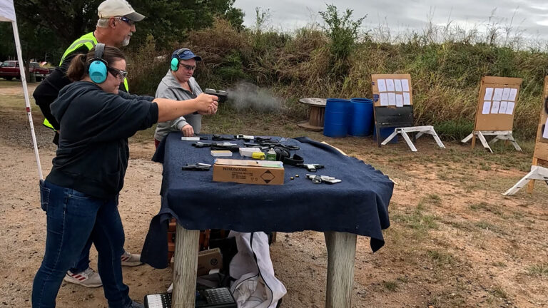 A participant fires a pistol downrange while a club instructor coaches from behind and another participant watches.