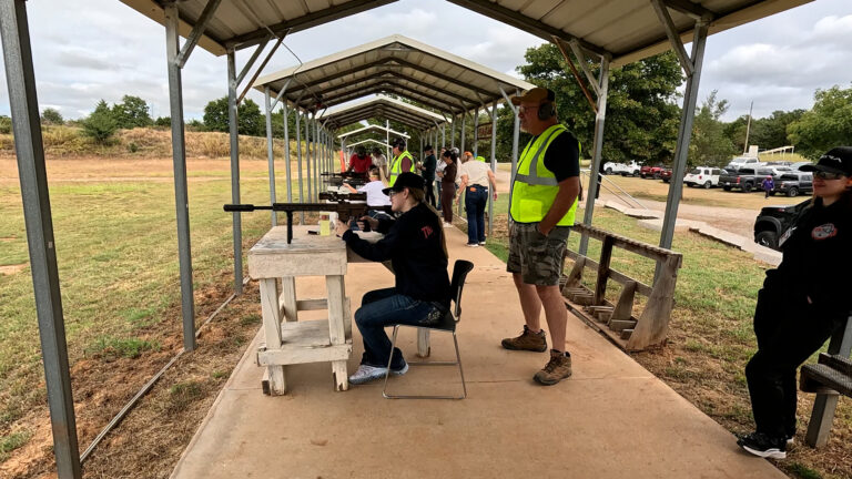 A participant fires a rifle from a bench on the club's covered long-range line while an instructor stands by.