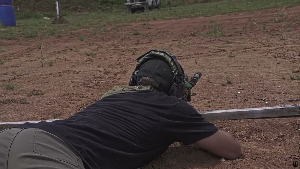 A shooter in the prone position fires a scoped rifle downrange toward the berm.
