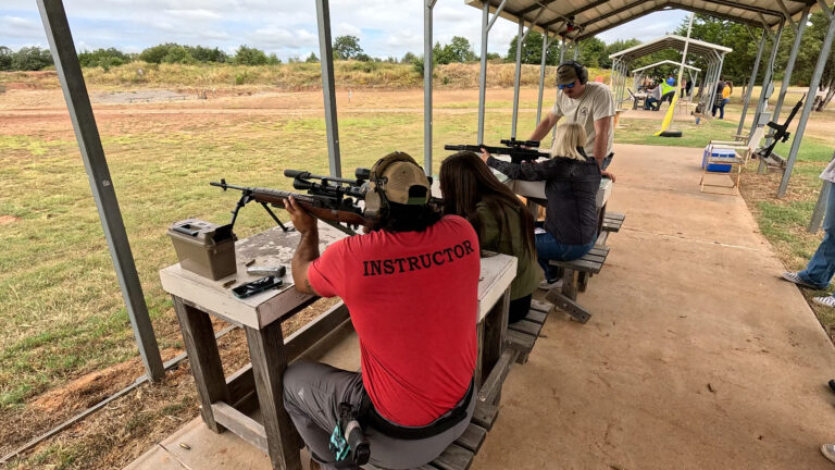 Several participants shoot rifles from the covered line while club instructors assist at each bench.