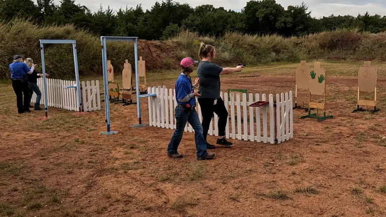 A participant works through a practical shooting course while a range officer walks alongside.