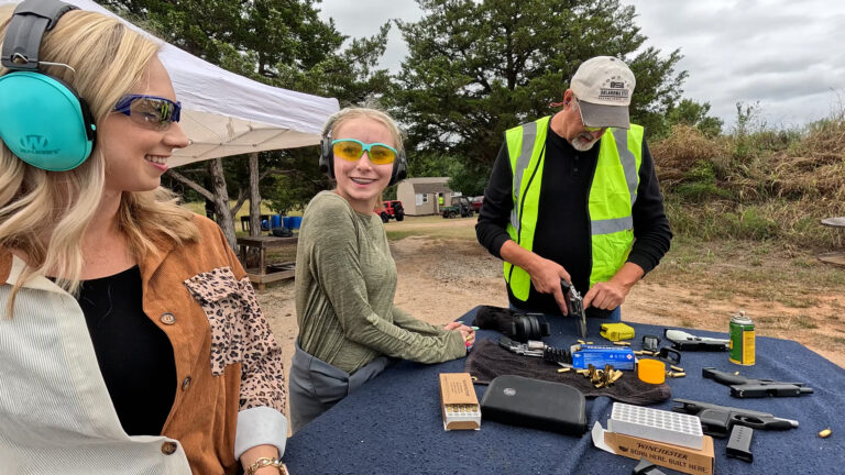 Two participants share a laugh at a shooting station while a club instructor prepares a revolver.